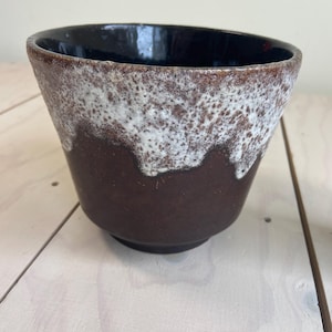 Pode incluir: Um vaso de cerâmica com base marrom escuro, transicionando para um meio salpicado branco e marrom, e um interior preto brilhante. O vaso tem um formato ligeiramente alargado.
