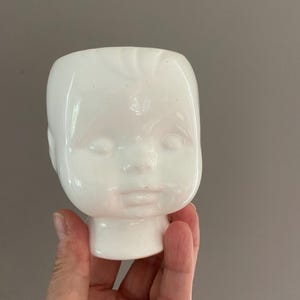 以下が含まれることがあります： デフォルメされた人の頭の形をした白い陶器製のプランター。滑らかで光沢のある仕上げで、閉じられた目、小さな鼻、唇などの顔の造形が施されています。頭頂部は開いており、中が空洞の容器になっています。