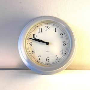 May include: A round, white quartz clock with a light gray frame. The clock face has black numerals and hands, with the time displayed as approximately 9:50. The word "QUARTZ" is printed at the bottom of the clock face.