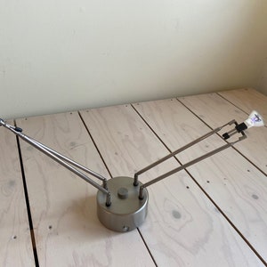 Può includere: Un apparecchio di illuminazione in metallo argentato con due bracci regolabili e due lampadine. L'apparecchio è montato su una base circolare.