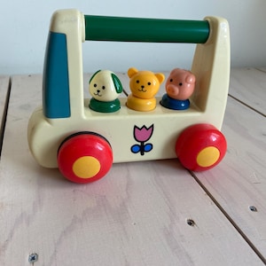 May include: A vintage toy bus with three animal figures: a dog, a bear, and a pig. The bus is cream-colored with red wheels and a green handle. A flower graphic is on the front. The toy is designed for young children.