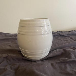 May include: A white ceramic planter with a barrel-shaped design, featuring horizontal ridges around the top and bottom sections. The vessel has a wide, open circular top and a rounded body that tapers slightly towards the base, with a smooth finish.