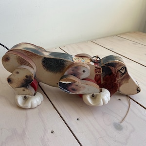 Op de afbeelding: Vintage houten trekdier hond met een bruin, zwart en wit gevlekt lichaam. De hond heeft een gedetailleerd geschilderd gezicht, rode wielen en een trekkoord. Een klassiek ontwerp, waarschijnlijk uit het midden van de 20e eeuw.