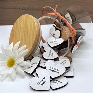 May include: A glass jar filled with small white wooden hearts, each with a different chore or task written on it. The jar is tipped over, spilling out the hearts onto a white surface. The hearts have phrases like "1 unfinished project", "1 book or magazine", "2 training items", and "1 bowl or plate".