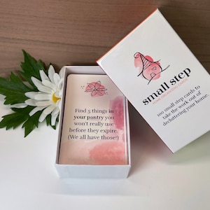May include: A white box with a pink and white card inside. The card has the text "Find 5 things in your pantry you won't really use before they expire. (We all have those!)". The box is open and the lid is on the right side of the image. The lid has the text "small step HOME ORGANIZING CARDS 100 small step cards to take the work out of decluttering your home."