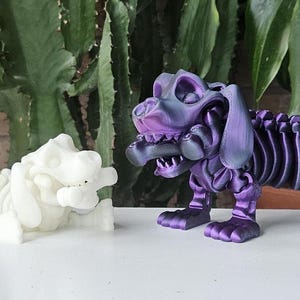 May include: Two 3D printed skeleton dog figurines. One is white and the other is a dark purple with a metallic sheen.