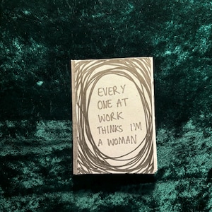 May include: A small, white notebook with black ink. The notebook has a circle drawn on the front with the text "EVERY ONE AT WORK THINKS I'M A WOMAN" inside.