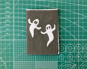 Pode incluir: Um pequeno caderno preto com duas ilustrações de fantasmas brancos na capa. Os fantasmas estão de frente um para o outro e têm traços simples e arredondados.