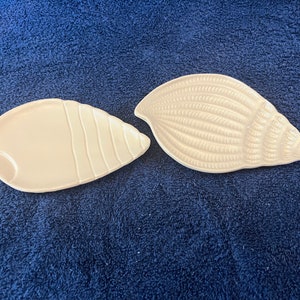 Puede incluir: Dos platos en forma de concha de color blanquecino. Uno tiene una superficie lisa con estrías verticales, el otro un diseño en espiral texturizado. Estos platos decorativos son ideales para servir aperitivos o como decoración costera.