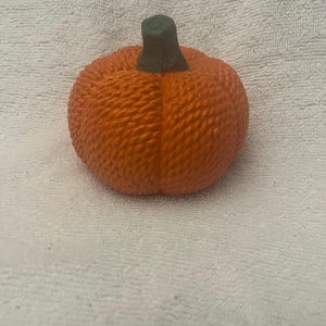 Op de afbeelding: Een kleine oranje pompoen met een groene steel. De pompoen is gemaakt van een geweven materiaal en heeft een textuur.