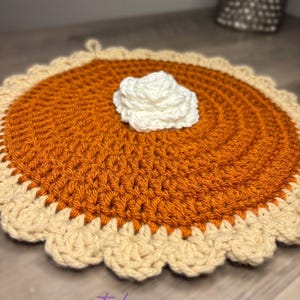 May include: A crocheted pumpkin pie-shaped trivet with a cream-colored whipped cream topping. The pie has an orange filling and a tan crust with a scalloped edge. The text "COME GET waisted" is visible in the bottom left corner.