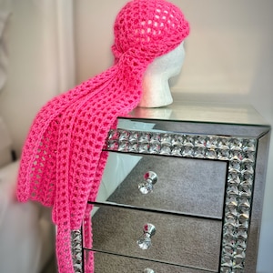 May include: A bright pink crocheted head wrap with a long tail. The wrap is on a mirrored nightstand with crystal accents. The nightstand has three drawers with crystal knobs. The text "COME GET waisted" is visible on the bottom drawer.