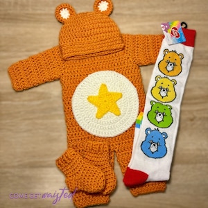 May include: A crocheted Care Bear costume for a baby, featuring a bright orange jumpsuit with a white star on the chest, a matching hat, and a pair of orange booties. The costume is paired with a white sock with a rainbow stripe and Care Bear characters printed on it.