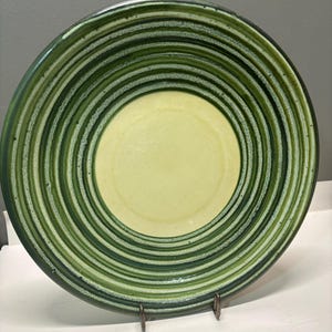 May include: A ceramic plate with a spiral design in shades of green and a yellow center. The plate is round and has a glossy finish. The design features concentric circles of varying shades of green, creating a textured appearance. The plate is displayed on a metal stand.
