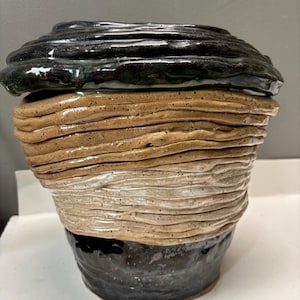 Puede incluir: Una maceta de cerámica hecha a mano con un diseño en capas. La parte superior es de un verde-negro oscuro y brillante, que pasa a bandas beige, blancas y negras. La superficie texturizada le da a esta pieza decorativa una sensación única y orgánica.