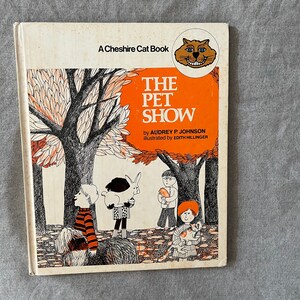 Puede incluir: Una portada de libro infantil vintage con un fondo naranja brillante. El título es "The Pet Show" con ilustraciones de niños y sus mascotas en un parque. El libro forma parte de la serie "A Cheshire Cat Book".