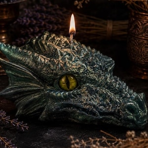 May include: A sculpted dragon head candle with a textured, dark green and metallic gold finish. The candle features a single lit wick on top and a prominent, realistic yellow eye with a vertical black pupil set into the side of the dragon's face.