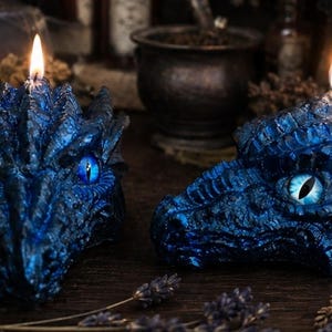 Peut inclure: Deux bougies sculptées en forme de tête de dragon avec une finition bleu métallique profond, des écailles texturées et des yeux bleus lumineux. Chaque bougie possède une mèche allumée au sommet, diffusant une lueur sur la surface détaillée.