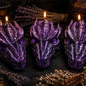 May include: Three purple dragon head candles with textured scales and glowing purple eyes. Each candle features a lit wick at the top of the head, casting a warm glow. The dragon heads are sculpted with intricate details, including ridges and horns.