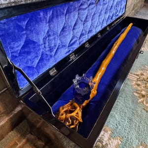 Op de afbeelding: Een houten toverstaf met een wortelachtige basis en een heldere glazen kroon, tentoongesteld in een open zwarte doos met blauwe fluwelen voering. De toverstaf is lichtbruin en rust op een blauw fluwelen kussen.
