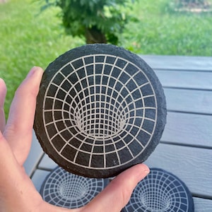 May include: A set of three black slate coasters with a white, grid-like design depicting a three-dimensional hole or vortex. The coasters are round and appear to be made of natural stone, with a rough, textured surface.
