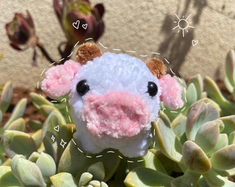 May include: A white and pink plush cow toy with brown ears and black eyes. The toy is sitting on a bed of green succulents.
