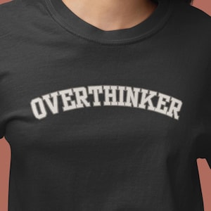 May include: A black short-sleeved crew neck t-shirt featuring the word OVERTHINKER printed in a white collegiate-style font across the chest. The garment has a standard fit with a ribbed neckline and straight hemline, constructed from a solid fabric.