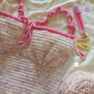 Op de afbeelding: Een gehaakt topje met een gespikkeld ontwerp in roze en wit. Het topje heeft een roze ruche-bies en een strik aan de voorkant. Het topje ligt op een wit stofoppervlak met een bol wol, haarclips en een flesje lipgloss.