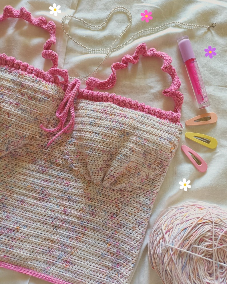 Op de afbeelding: Een gehaakt topje met een gespikkeld ontwerp in roze en wit. Het topje heeft een roze ruche-bies en een strik aan de voorkant. Het topje ligt op een wit stofoppervlak met een bol wol, haarclips en een flesje lipgloss.