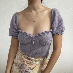Op de afbeelding: Een lavendelkleurig gehaakt topje met een sweetheart-halslijn en korte pofmouwen. Het topje is gecropped en heeft een delicaat strikdetail in het midden.