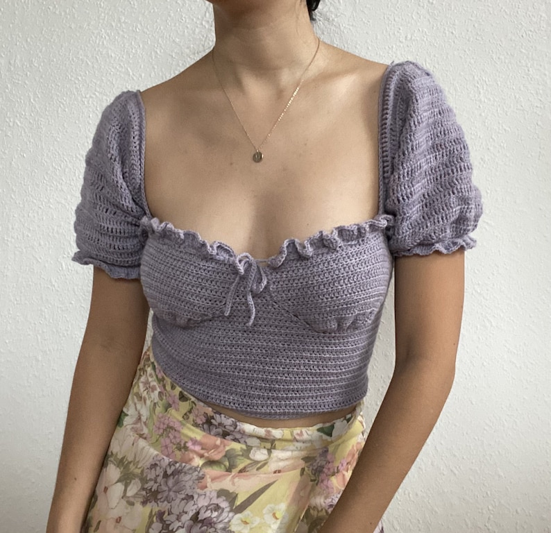 Op de afbeelding: Een lavendelkleurig gehaakt topje met een sweetheart-halslijn en korte pofmouwen. Het topje is gecropped en heeft een delicaat strikdetail in het midden.