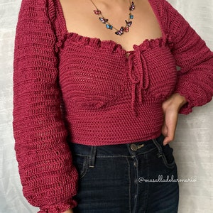 Op de afbeelding: Een close-up van een vrouw die een kastanjebruin gecrochete top draagt met een vlinderketting. De top heeft een vierkante halslijn en lange mouwen met ruches aan de manchetten. De vrouw draagt ook donkerblauwe jeans.