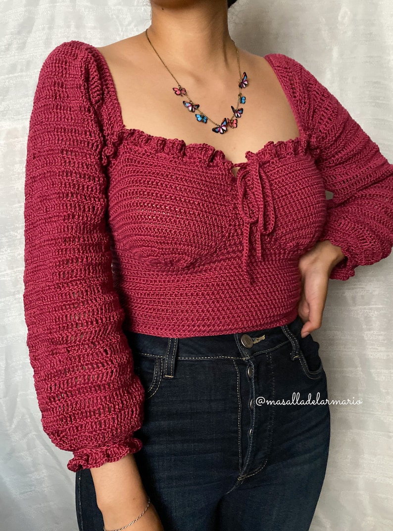 Op de afbeelding: Een close-up van een vrouw die een kastanjebruin gecrochete top draagt met een vlinderketting. De top heeft een vierkante halslijn en lange mouwen met ruches aan de manchetten. De vrouw draagt ook donkerblauwe jeans.