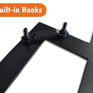 May include: Close-up of a black metal sign with two built-in hooks. The hooks are attached to the sign with screws and have a serrated edge.