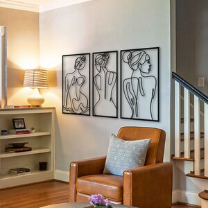 May include: Three black metal wall sculptures depicting stylised figures of women. The sculptures are arranged in a row on a white wall. The sculptures are in a living room setting with a brown leather armchair and a white bookshelf.