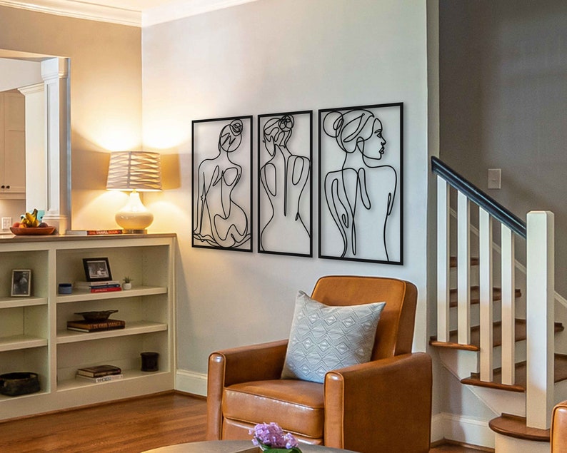 May include: Three black metal wall sculptures depicting stylised figures of women. The sculptures are arranged in a row on a white wall. The sculptures are in a living room setting with a brown leather armchair and a white bookshelf.