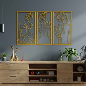 May include: Three yellow metal wall art pieces featuring line drawings of women's figures. The figures are stylised and abstract, with flowing lines and curves. The art pieces are hung on a grey wall above a wooden shelf with decorative items.