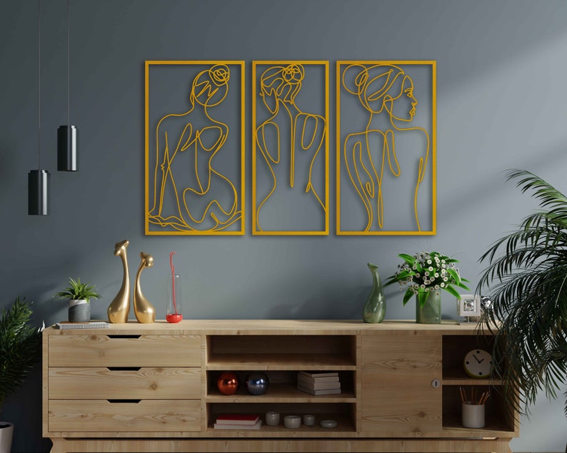 May include: Three yellow metal wall art pieces featuring line drawings of women's figures. The figures are stylised and abstract, with flowing lines and curves. The art pieces are hung on a grey wall above a wooden shelf with decorative items.