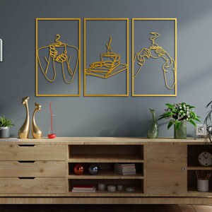 May include: Three gold metal wall art pieces featuring a line art design of people holding coffee cups. The art is hanging on a grey wall above a wooden shelf with decorative items.