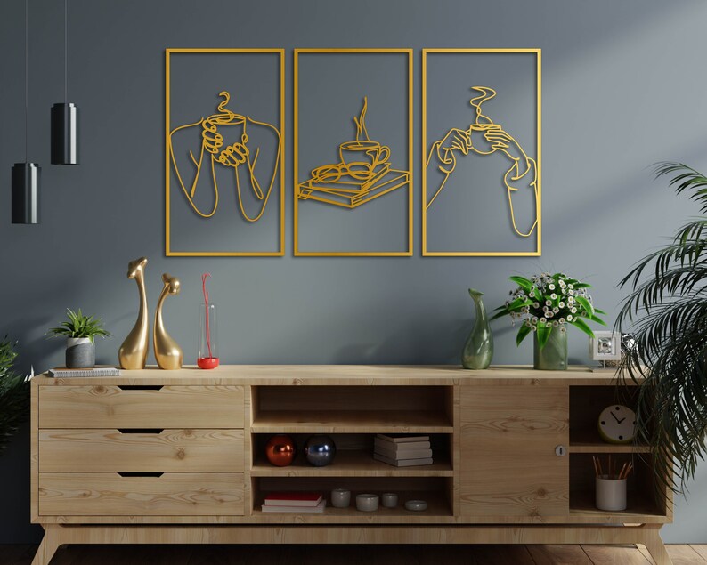 May include: Three gold metal wall art pieces featuring a line art design of people holding coffee cups. The art is hanging on a grey wall above a wooden shelf with decorative items.