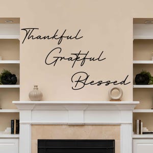 Könnte beinhalten: Ein schwarzer Wandaufkleber mit den Wörtern Thankful, Grateful und Blessed in einer kursiven Schriftart. Die Wörter sind in drei separaten Zeilen untereinander auf einer hellbeigen Wand über einem weißen Kamin angeordnet.