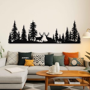 Può includere: Arte da parete in metallo nero raffigurante una scena di foresta con pini sagomati e due cervi. L'opera d'arte è montata su una parete bianca sopra un divano grigio con cuscini decorativi e un tavolino in legno.