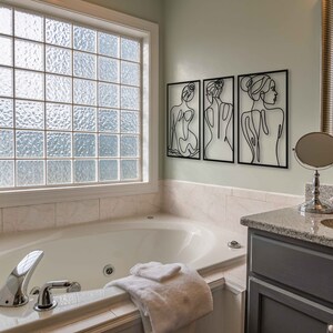 May include: A white bathtub with chrome fixtures and a white towel in a bathroom with a window, a vanity with a sink, and three black and white line art prints of women on the wall.