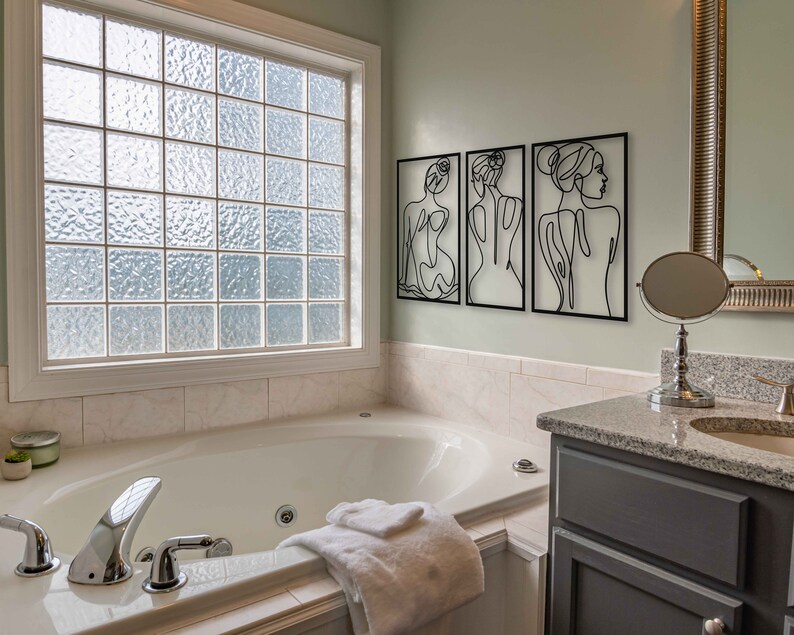 May include: A white bathtub with chrome fixtures and a white towel in a bathroom with a window, a vanity with a sink, and three black and white line art prints of women on the wall.