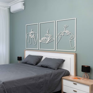 May include: Three white metal wall art pieces with line drawings of a person holding a cup of coffee, a cup of coffee with a plate of food, and a person holding a cup of coffee. The art is hanging on a light green wall above a grey bed with white bedding and a bedside table.