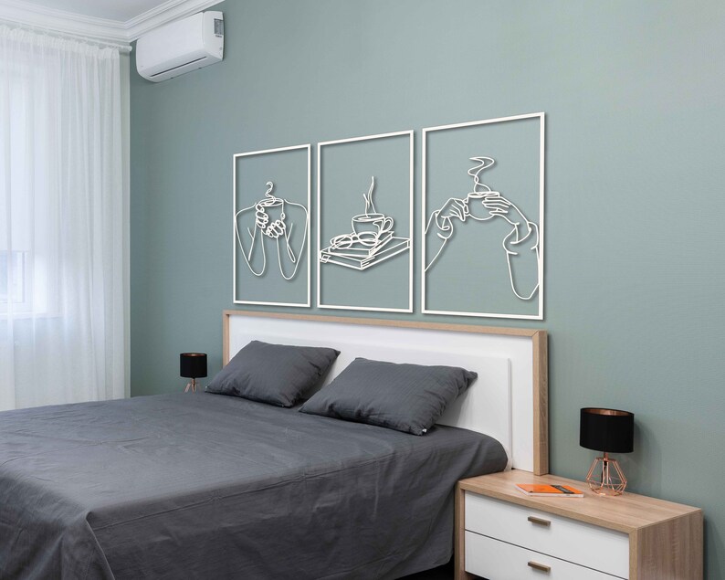 May include: Three white metal wall art pieces with line drawings of a person holding a cup of coffee, a cup of coffee with a plate of food, and a person holding a cup of coffee. The art is hanging on a light green wall above a grey bed with white bedding and a bedside table.