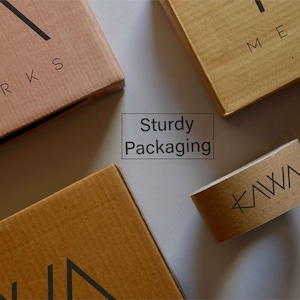 May include: Three brown cardboard boxes with the text "KAIWA WORKS" printed on them. The boxes are on a white surface. One box has the text "META" printed on it. The text "Sturdy Packaging" is printed on a white rectangle in the centre of the image.