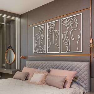 May include: Three white metal wall art pieces featuring line drawings of female figures. The art is hung on a grey wall above a bed with a grey headboard and white bedding.