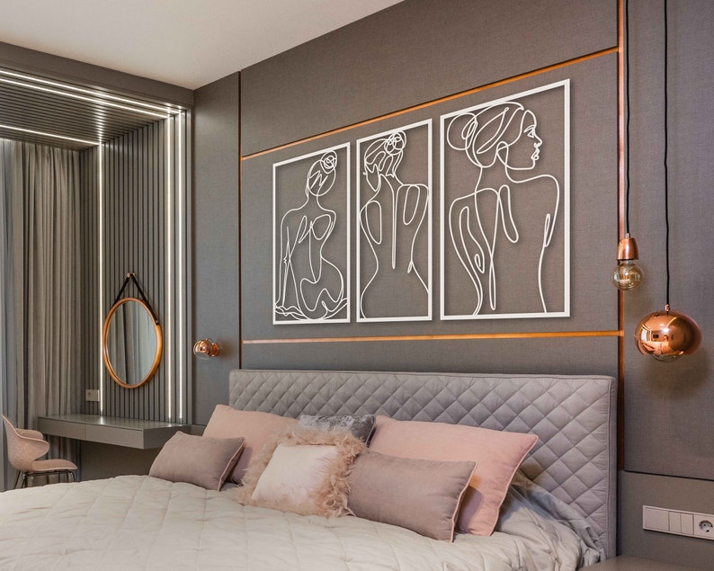 May include: Three white metal wall art pieces featuring line drawings of female figures. The art is hung on a grey wall above a bed with a grey headboard and white bedding.