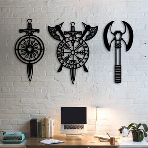 Pode incluir: Três peças de arte de parede em metal preto com designs de inspiração nórdica. A peça da esquerda é uma espada e um símbolo solar circular. A peça central mostra uma bússola Vegvisir atrás de dois machados cruzados e uma espada.
